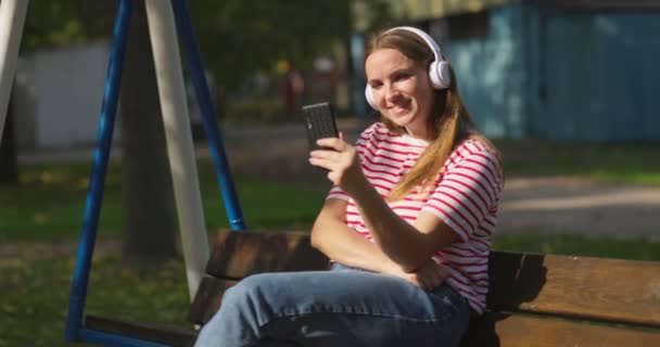 スイングに揺れるワイヤレスヘッドフォンで若い女性は 都市公園で音楽を聴く 高品質4K映像 — ストック動画
