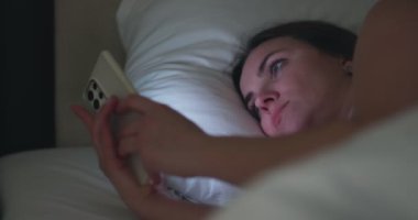 A woman lies in bed at night and uses a smartphone. Female using mobile phone browsing social media, chatting, dating apps, doing internet online shopping before bedtime. High quality 4k footage