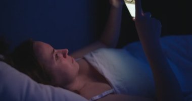 A woman lies in bed at night and uses a smartphone. Female using mobile phone browsing social media, chatting, dating apps, doing internet online shopping before bedtime. High quality 4k footage