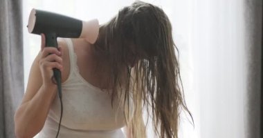 Beautiful young woman using hair dryer near the window at home. High quality 4k footage