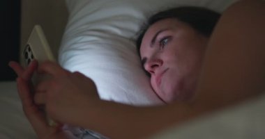 A woman lies in bed at night and uses a smartphone. Female using mobile phone browsing social media, chatting, dating apps, doing internet online shopping before bedtime. High quality 4k footage