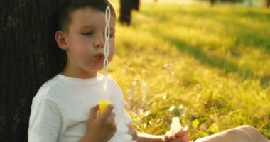 Child boy blows soap bubbles, having fun playing in park in rays of sunset. Child plays with soap bubbles in the summer garden. Happy childhood concept. High quality 4k footage