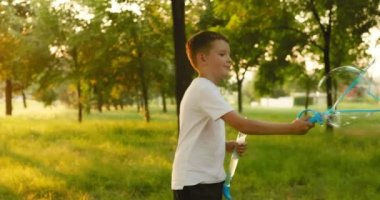 Child boy blows soap bubbles, having fun playing in park in rays of sunset. Child plays with soap bubbles in the summer garden. Happy childhood concept. High quality 4k footage