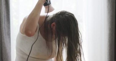 Beautiful young woman using hair dryer near the window at home. High quality 4k footage