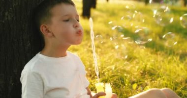 Child boy blows soap bubbles, having fun playing in park in rays of sunset. Child plays with soap bubbles in the summer garden. Happy childhood concept. High quality 4k footage