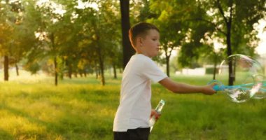 Child boy blows soap bubbles, having fun playing in park in rays of sunset. Child plays with soap bubbles in the summer garden. Happy childhood concept. High quality 4k footage