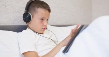 Beyaz tişörtlü, kulaklıklı tablet bilgisayar kullanan, evde yatağında oturan bir çocuk. Yüksek kalite 4k görüntü