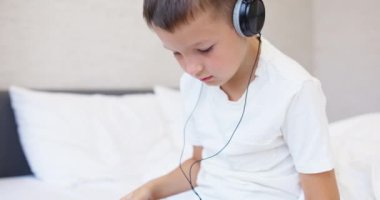 Beyaz tişörtlü, kulaklıklı tablet bilgisayar kullanan, evde yatağında oturan bir çocuk. Yüksek kalite 4k görüntü