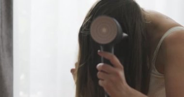 Evdeki pencerenin yanında saç kurutma makinesi kullanan güzel bir genç kadın. Yüksek kalite 4k görüntü