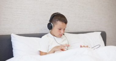 Beyaz tişörtlü, kulaklıklı tablet bilgisayar kullanan, evde yatağında oturan bir çocuk. Yüksek kalite 4k görüntü
