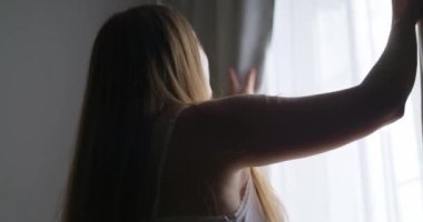 Güzel, genç esmer kadın, sabah güneşinin rahat dairesini aydınlatması için perdeleri açıyor. Kadın perdeleri açıyor ve gün ışığının tadını çıkarıyor. Yüksek kalite 4k görüntü