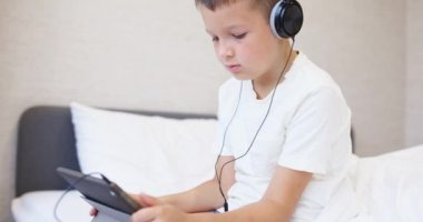 Beyaz tişörtlü, kulaklıklı tablet bilgisayar kullanan, evde yatağında oturan bir çocuk. Yüksek kalite 4k görüntü