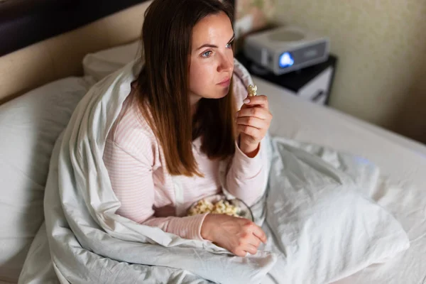 年轻女人正在家里看放映机 看电视 看电影 还在床上吃爆米花 — 图库照片