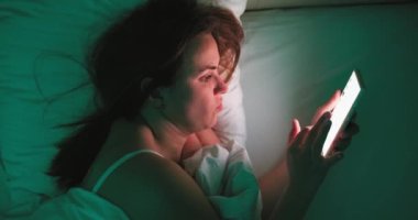 A woman lies in bed at night and uses a smartphone. Female using mobile phone browsing social media, chatting, dating apps, doing internet online shopping before bedtime. High quality 4k footage