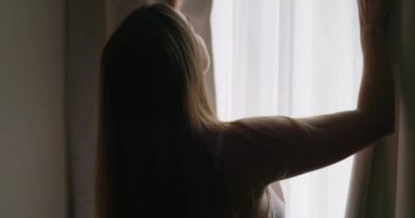 Güzel, genç esmer kadın, sabah güneşinin rahat dairesini aydınlatması için perdeleri açıyor. Kadın perdeleri açıyor ve gün ışığının tadını çıkarıyor. Yüksek kalite 4k görüntü