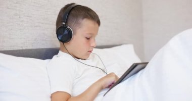 Beyaz tişörtlü, kulaklıklı tablet bilgisayar kullanan, evde yatağında oturan bir çocuk. Yüksek kalite 4k görüntü
