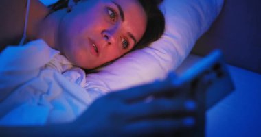 A woman lies in bed at night and uses a smartphone. Female using mobile phone browsing social media, chatting, dating apps, doing internet online shopping before bedtime. High quality 4k footage