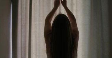 Güzel, genç esmer kadın, sabah güneşinin rahat dairesini aydınlatması için perdeleri açıyor. Kadın perdeleri açıyor ve gün ışığının tadını çıkarıyor. Yüksek kalite 4k görüntü