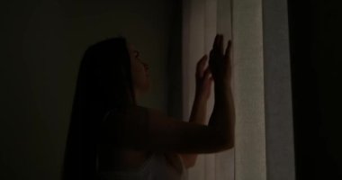 Güzel, genç esmer kadın, sabah güneşinin rahat dairesini aydınlatması için perdeleri açıyor. Kadın perdeleri açıyor ve gün ışığının tadını çıkarıyor. Yüksek kalite 4k görüntü
