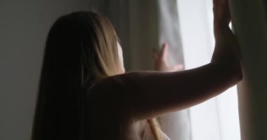 Güzel, genç esmer kadın, sabah güneşinin rahat dairesini aydınlatması için perdeleri açıyor. Kadın perdeleri açıyor ve gün ışığının tadını çıkarıyor. Yüksek kalite 4k görüntü