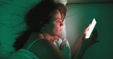 A woman lies in bed at night and uses a smartphone. Female using mobile phone browsing social media, chatting, dating apps, doing internet online shopping before bedtime. High quality 4k footage