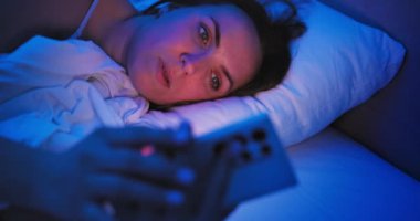 Bir kadın gece yatağa uzanıyor ve akıllı telefon kullanıyor. Cep telefonu kullanan, sosyal medyada gezinen, sohbet eden, flört uygulamaları kullanan, yatmadan önce internet alışverişi yapan bir kadın. Yüksek kalite 4k görüntü