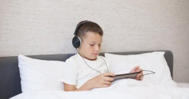 Beyaz tişörtlü, kulaklıklı tablet bilgisayar kullanan, evde yatağında oturan bir çocuk. Yüksek kalite 4k görüntü