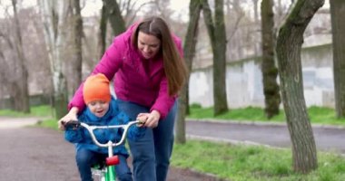 Anne oğluna bisiklet sürmeyi öğretiyor. Genç anne oğluna ilk kez parkta bisiklet sürmeyi öğretiyor. Bisikletli mutlu çocuklar. Birlikte geçirdiğimiz en güzel anlar. Çocukluk bisikleti konsepti. Yüksek kalite 4k görüntü