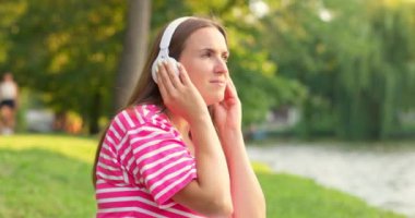 Çimenlerde oturmuş, parkta kulaklıkla müzik dinleyen rahat bir kadın. Yüksek kalite 4k görüntü