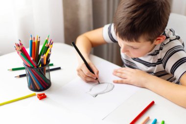 Bir çocuk masada otururken renkli kalemlerle beyaz kağıda resim çizer..