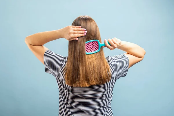 Back view of young brunette woman with hair brush brushing her long healthy hair on color background.