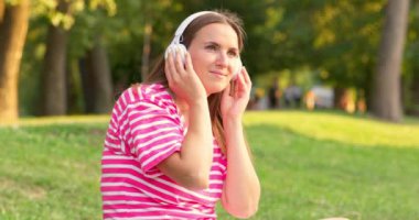 Çimenlerde oturmuş, parkta kulaklıkla müzik dinleyen rahat bir kadın. Yüksek kalite 4k görüntü