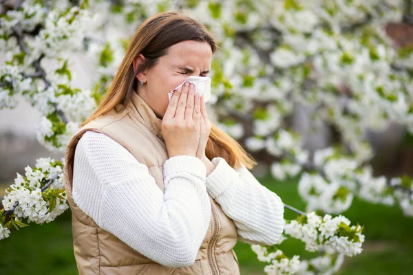 Sneezing woman suffering from seasonal allergy at spring with a nose wiper among the flowering trees in the park. Concept of spring allergies