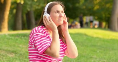 Çimenlerde oturmuş, parkta kulaklıkla müzik dinleyen rahat bir kadın. Yüksek kalite 4k görüntü