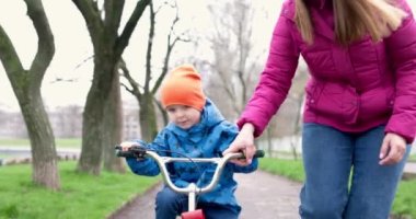 Anne oğluna bisiklet sürmeyi öğretiyor. Genç anne oğluna ilk kez parkta bisiklet sürmeyi öğretiyor. Bisikletli mutlu çocuklar. Birlikte geçirdiğimiz en güzel anlar. Çocukluk bisikleti konsepti. Yüksek kalite 4k görüntü
