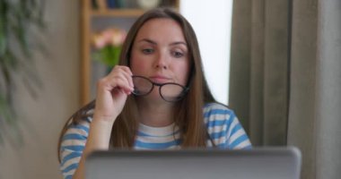 Gözlüklü düşünceli bir kadın dizüstü bilgisayarda çalışıyor, başka tarafa bakıyor, ev ofisindeki bir soruna çözüm bulmaya çalışıyor, karar veriyor, fikir yoksunluğu yaşıyor, manzarayı kapatıyor.