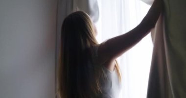 Güzel, genç esmer kadın, sabah güneşinin rahat dairesini aydınlatması için perdeleri açıyor. Kadın perdeleri açıyor ve gün ışığının tadını çıkarıyor. Yüksek kalite 4k görüntü