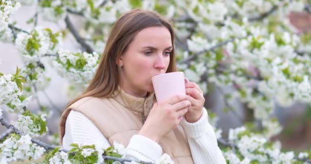 新鮮な香りのお茶を飲んでいる女性は 花の木のそばに立って味を楽しんでいます 高品質4K映像 — ストック動画