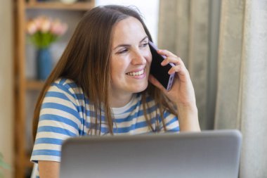 Bağlantı ve iletişim. Evde çalışan gülümseyen bir kadın cep telefonuyla konuşan bir dizüstü bilgisayar kullanıyor. Mesafe serbest meslek kavramı