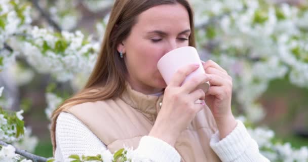 新鮮な香りのお茶を飲んでいる女性は 花の木のそばに立って味を楽しんでいます 高品質4K映像 — ストック動画