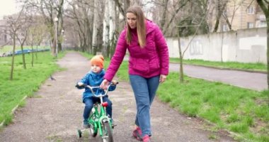 Anne oğluna bisiklet sürmeyi öğretiyor. Genç anne oğluna ilk kez parkta bisiklet sürmeyi öğretiyor. Bisikletli mutlu çocuklar. Birlikte geçirdiğimiz en güzel anlar. Çocukluk bisikleti konsepti. Yüksek kalite 4k görüntü