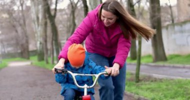 Anne oğluna bisiklet sürmeyi öğretiyor. Genç anne oğluna ilk kez parkta bisiklet sürmeyi öğretiyor. Bisikletli mutlu çocuklar. Birlikte geçirdiğimiz en güzel anlar. Çocukluk bisikleti konsepti. Yüksek kalite 4k görüntü