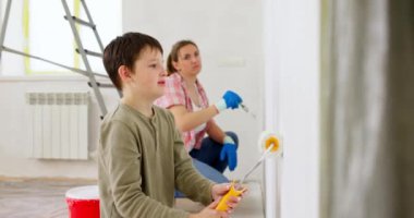 Anne ve oğlu boya silindiri ve fırçayla duvar boyuyor, birlikte tadilatın keyfini çıkarıyorlar. Ailecek vakit geçireceğiz. Kalacak yer ve aile kavramı. Yüksek kalite 4k görüntü