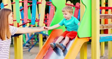 Mutlu anne ve küçük oğlu oyun parkı kaydırağında oynuyorlar ve parkta eğleniyorlar. Çocuklar için sağlıklı bir yaz aktivitesi. Yüksek kalite 4k görüntü