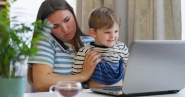 Anne kucağında oğluyla uzaktan kumandayla evden bilgisayarda çalışıyor. Anne evde çalışmaya çalışıyor, çocuk da ellerinin üstünde oturuyor. Yüksek kalite 4k görüntü