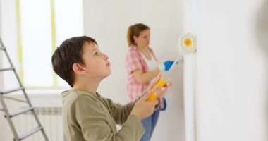 Anne ve oğlu boya silindiri ve fırçayla duvar boyuyor, birlikte tadilatın keyfini çıkarıyorlar. Ailecek vakit geçireceğiz. Kalacak yer ve aile kavramı. Yüksek kalite 4k görüntü