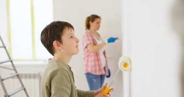 Anne ve oğlu boya silindiri ve fırçayla duvar boyuyor, birlikte tadilatın keyfini çıkarıyorlar. Ailecek vakit geçireceğiz. Kalacak yer ve aile kavramı. Yüksek kalite 4k görüntü