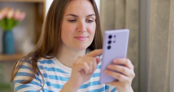 スマートフォンを持っている幸せな女性は 彼女の携帯電話の画面を見て モバイルショッピングアプリを楽しんで 楽しいゲームをしたり ソーシャルメディアでチャットしたり 自宅に座っています 高品質4K映像 — ストック動画