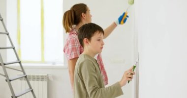 Anne ve oğlu boya silindiri ve fırçayla duvar boyuyor, birlikte tadilatın keyfini çıkarıyorlar. Ailecek vakit geçireceğiz. Kalacak yer ve aile kavramı. Yüksek kalite 4k görüntü