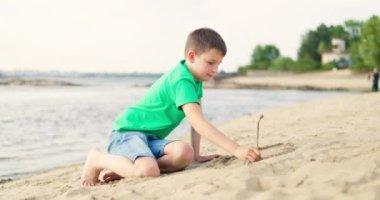 Çocuk yazın sıcak güneş ışığında kumsalda kumla oynuyor. Çocuk kumda çizim yapıyor. Yaz turizmi ve eğlence konsepti. Yüksek kalite 4k görüntü